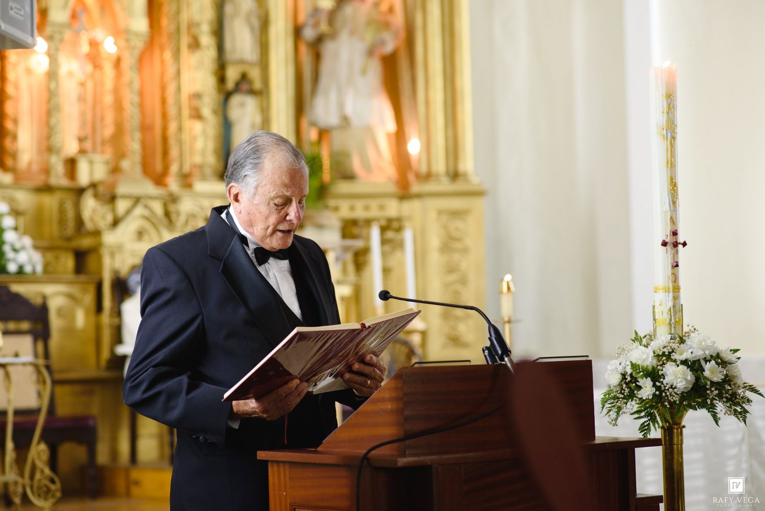 Elderly man in tuxedo reading from book at wooden podium inside ornate Catholic church during wedding ceremony.