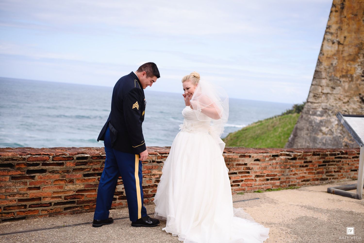 Military groom and blonde bride share a tender moment at a coastal brick fort overlook on their wedding day.