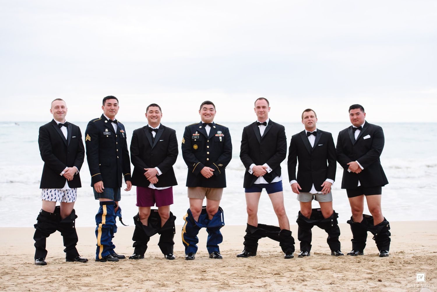 Groomsmen in military uniforms and tuxedos pose humorously without pants on a sandy beach.