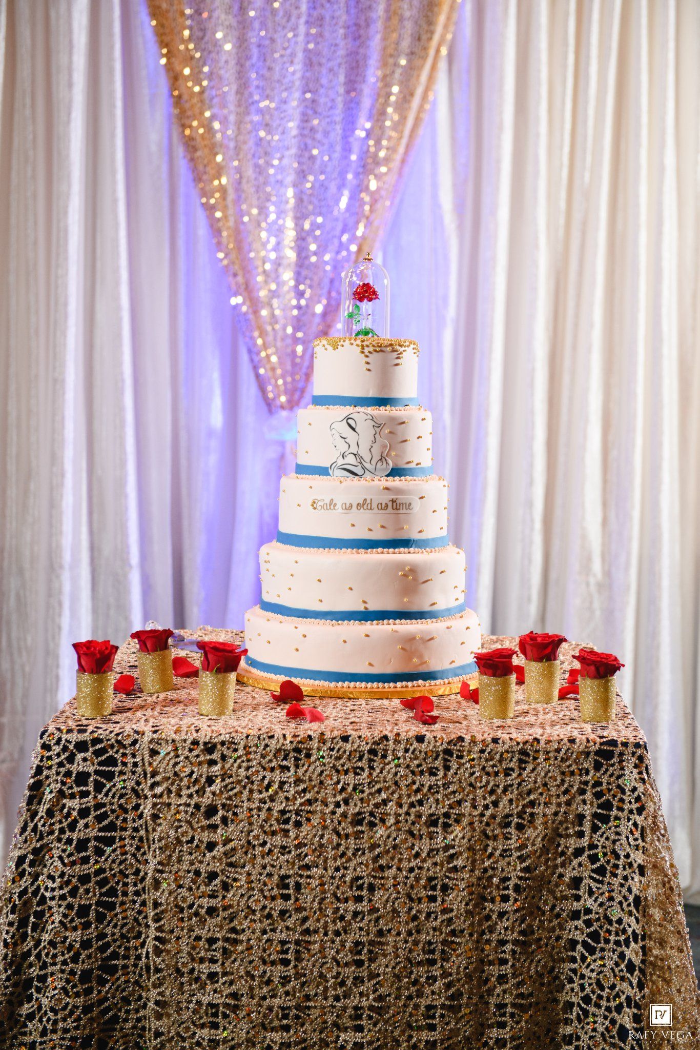Elegant five-tier white wedding cake with blue accents and red roses on a decorative table with sparkling backdrop.