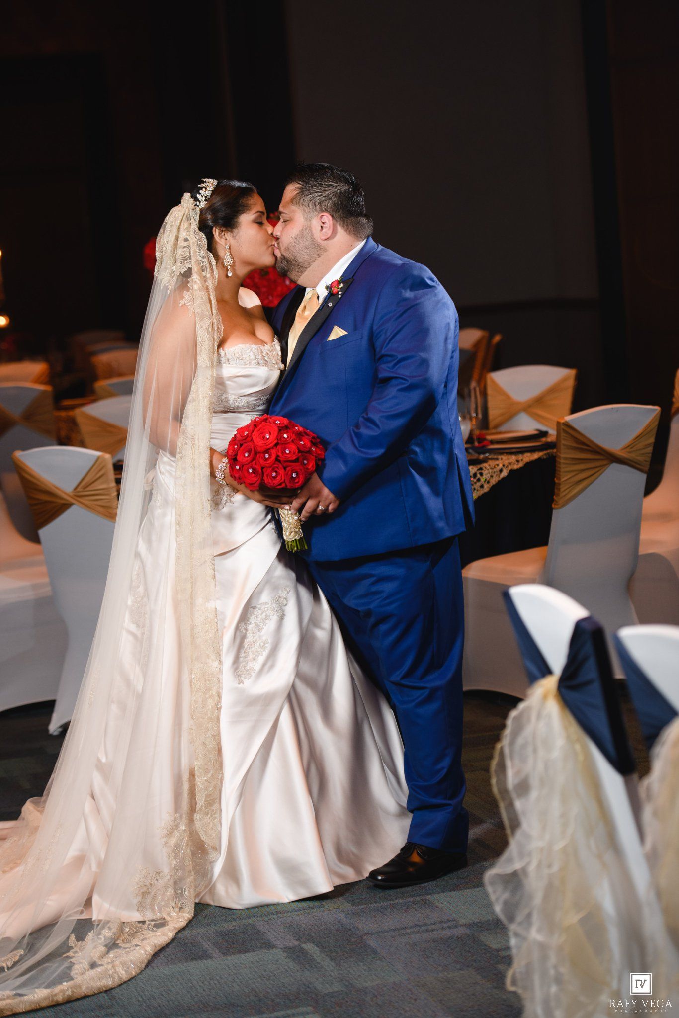 Bride and groom share first kiss at wedding ceremony, bride in white gown with veil holding red rose bouquet.
