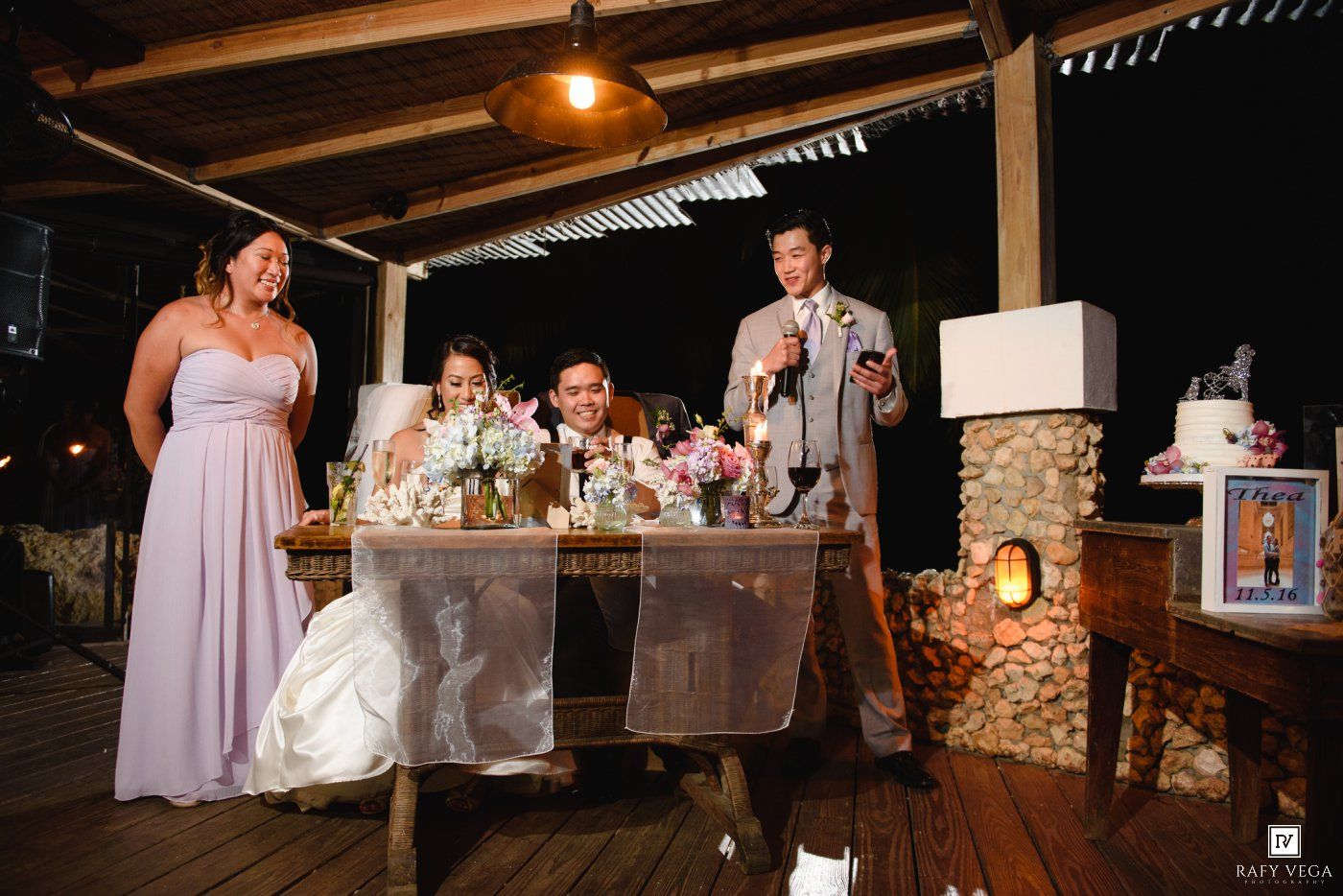 Groom gives a toast at wedding reception sweetheart table with bride and bridesmaid smiling nearby.