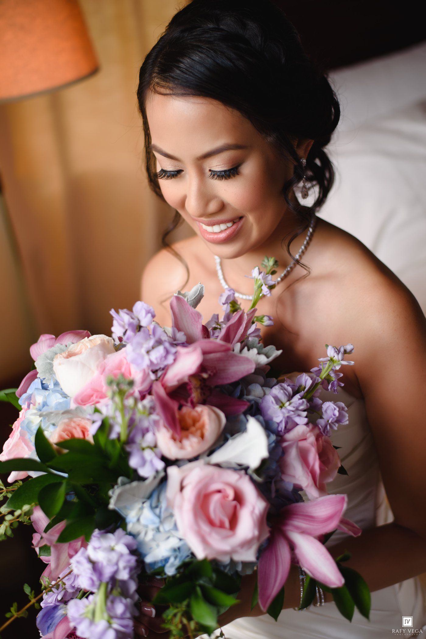 A radiant bride smiles while holding a lush bouquet of pink roses, lilies, and purple flowers on her wedding day.