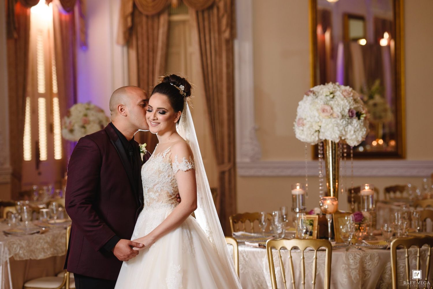 Bride in white gown and groom in burgundy suit share romantic moment at elegant gold wedding reception.