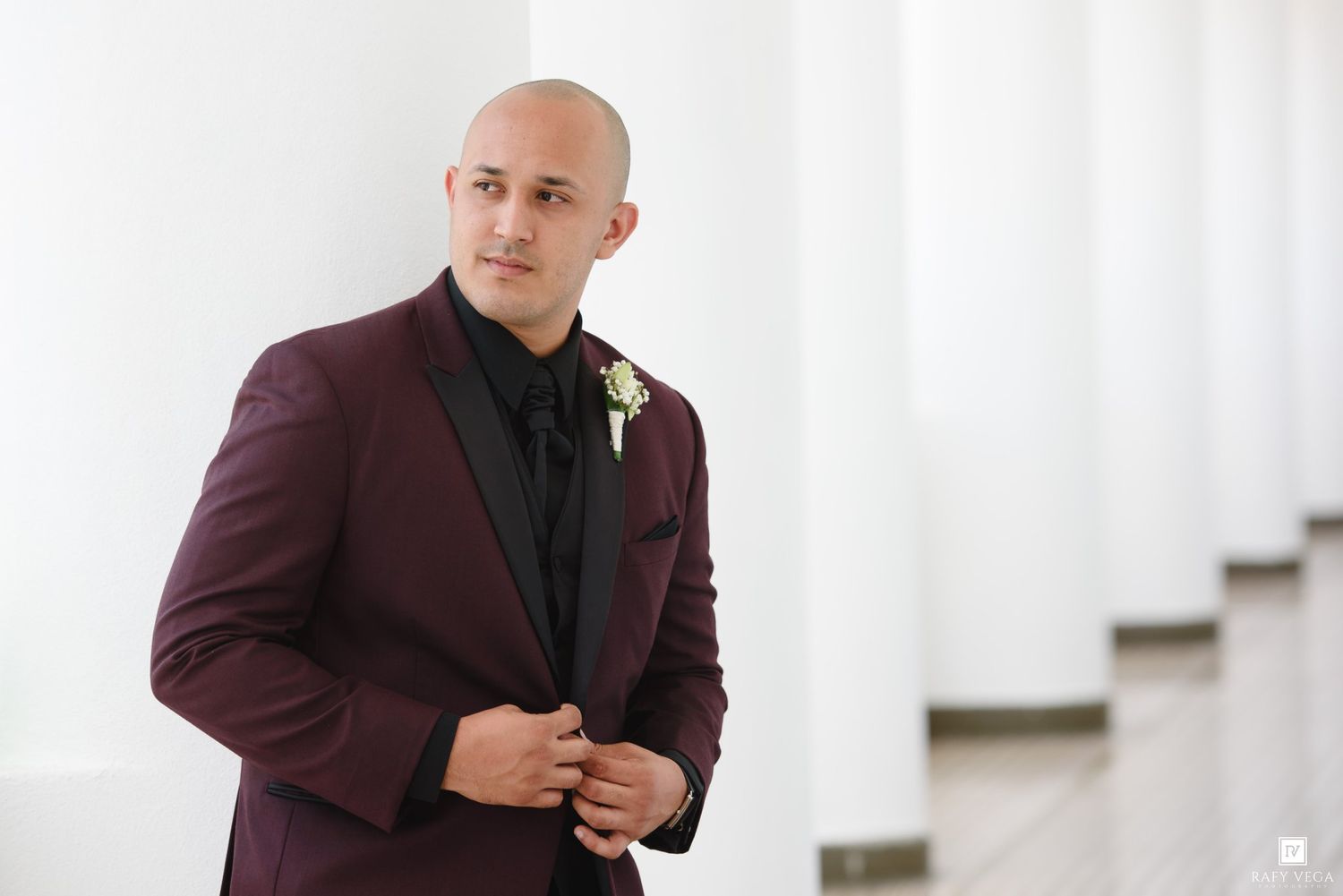 Bald groom in burgundy tuxedo with black lapels and boutonniere poses confidently against white columns.