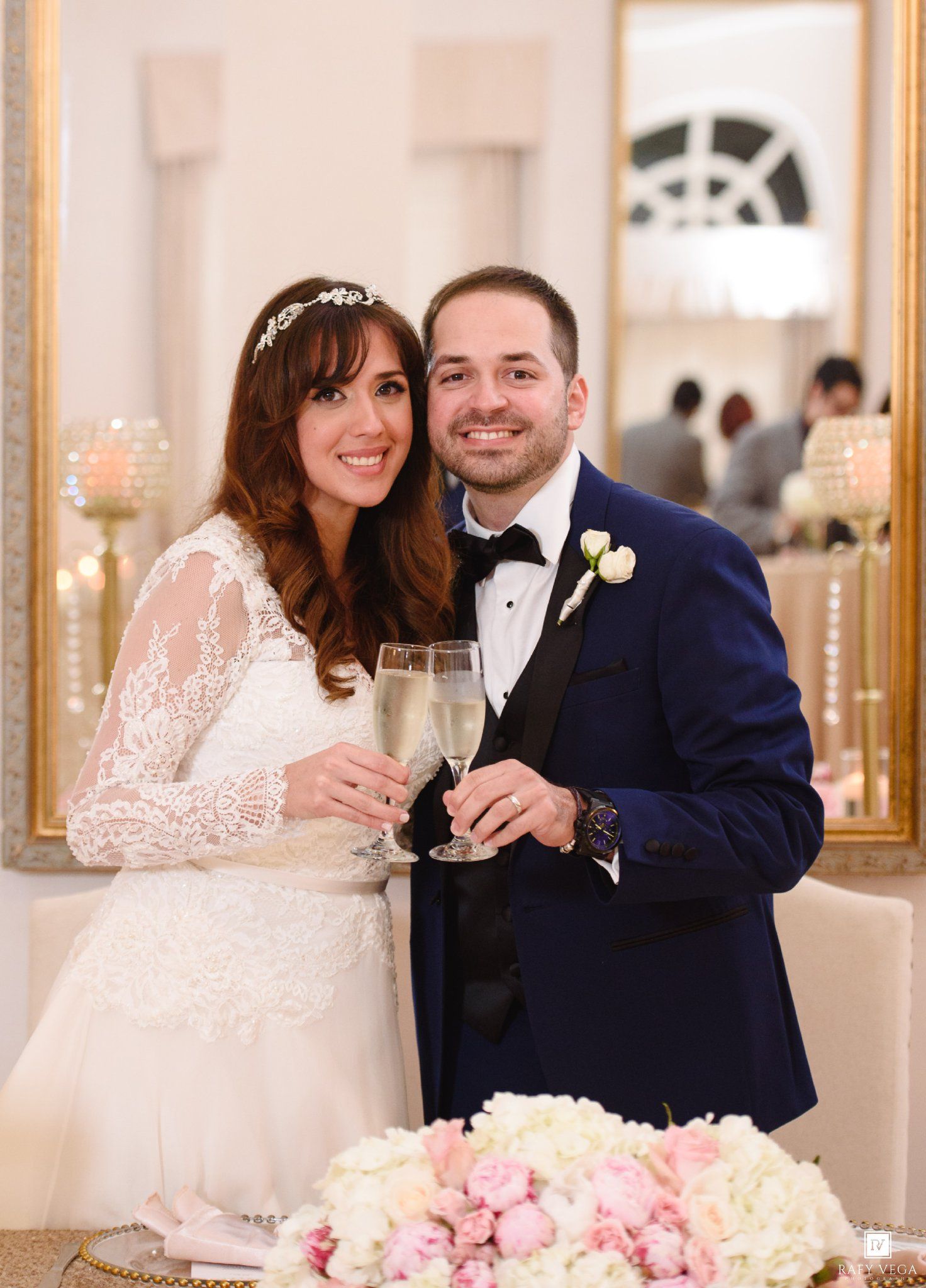Bride and groom toasting with champagne glasses at their wedding reception, wearing white lace gown and navy tuxedo.