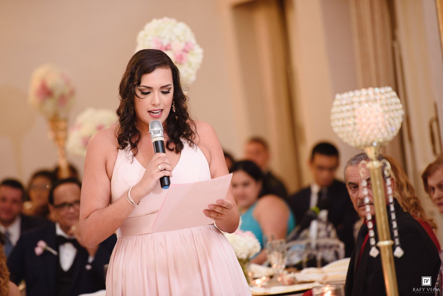 Bridesmaid in pink gown delivers heartfelt speech at elegant wedding reception, holding microphone and paper.