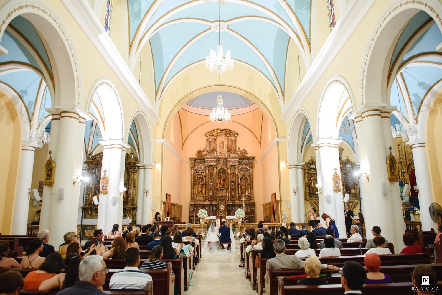 Elegant church wedding ceremony with guests seated in wooden pews beneath a stunning blue vaulted ceiling and gilded altar.