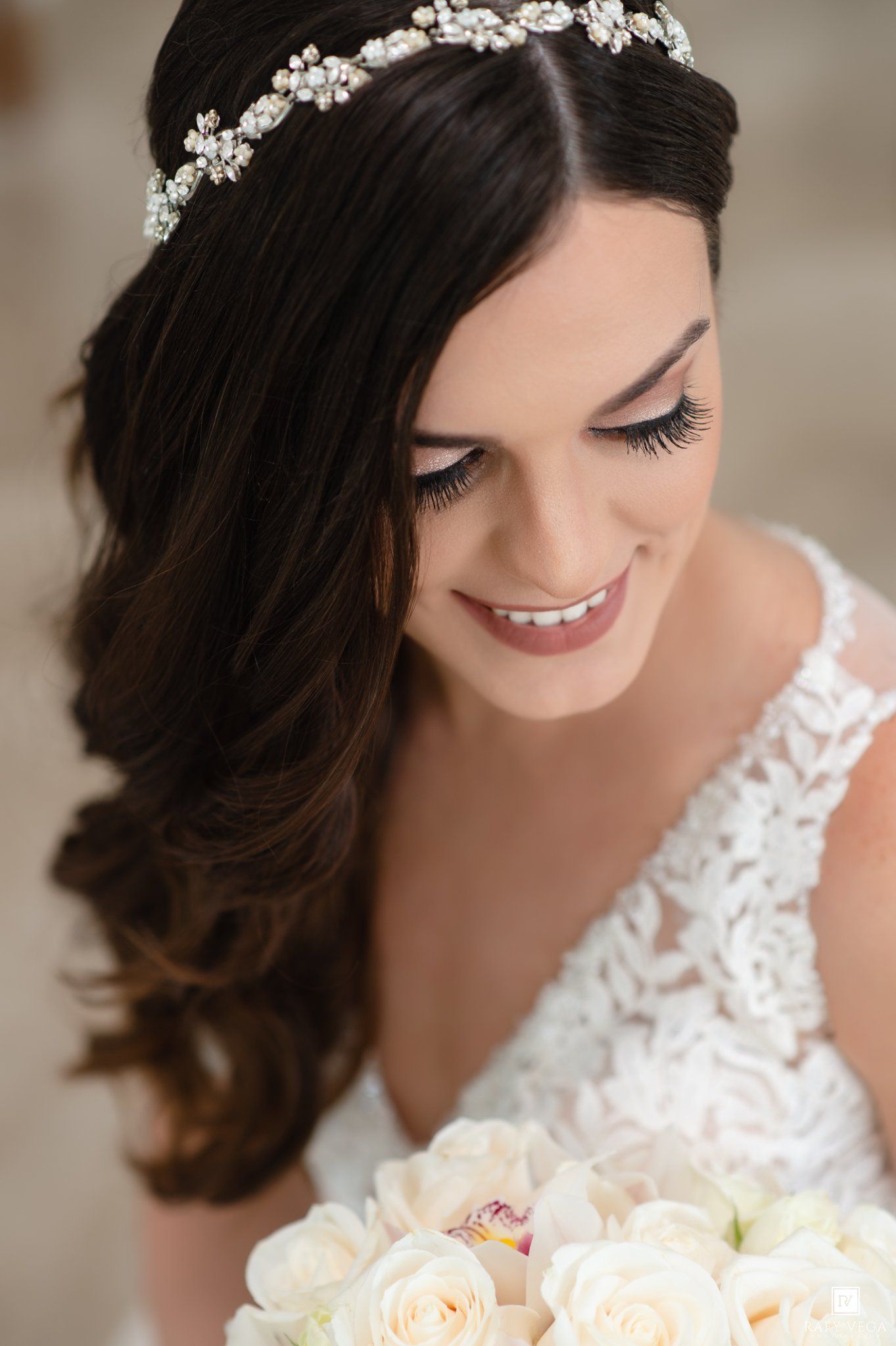 Smiling bride with dark hair, crystal headpiece, lace gown, and white rose bouquet on her wedding day.
