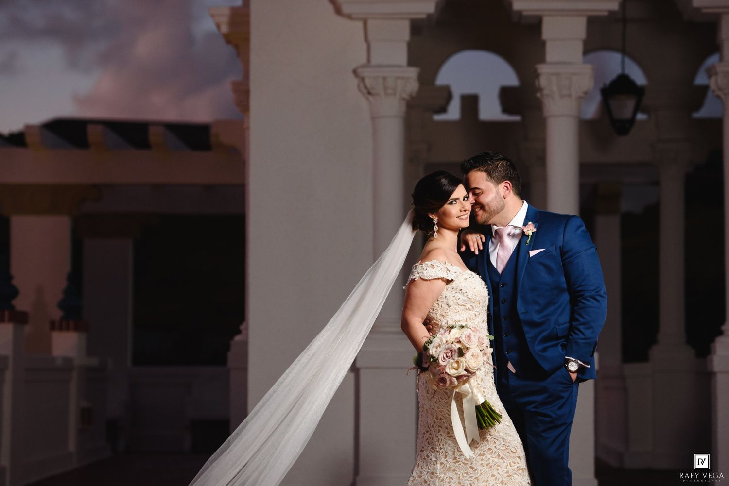 Bride and groom share romantic moment at sunset on elegant venue balcony, bride in lace gown with flowing veil.
