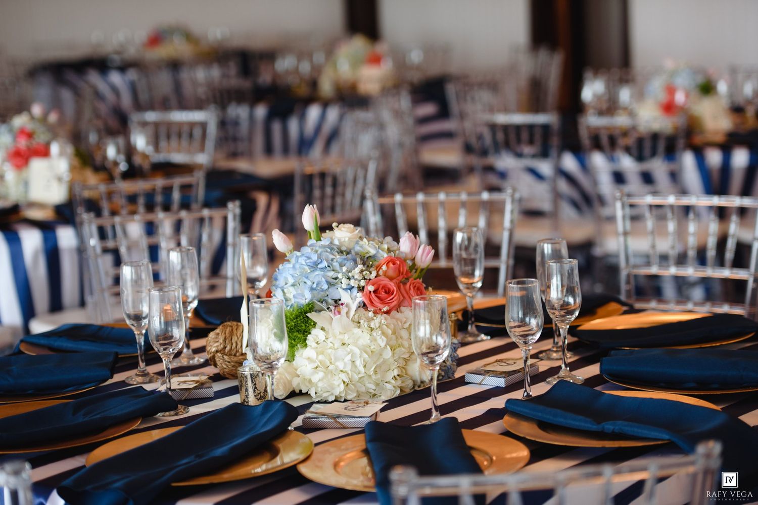 Elegant wedding reception tables with navy blue linens, floral centerpieces of white hydrangeas and coral roses, and crystal glassware.