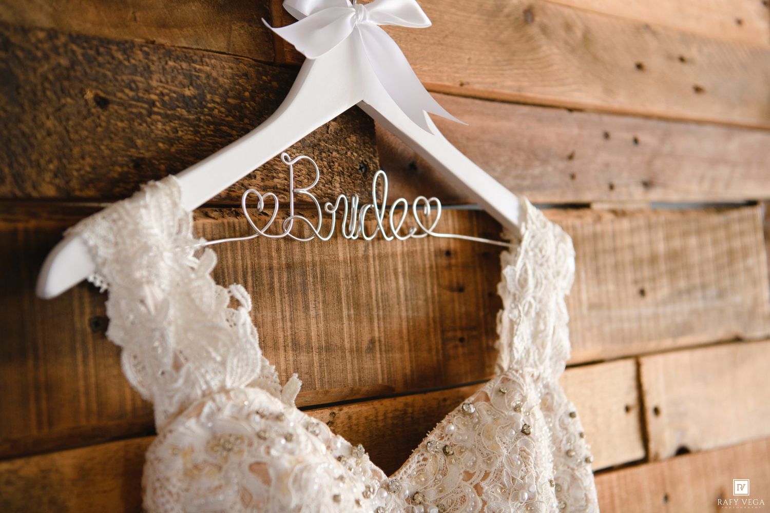 Lace wedding dress hanging on a white Bride hanger against a rustic wooden wall backdrop.
