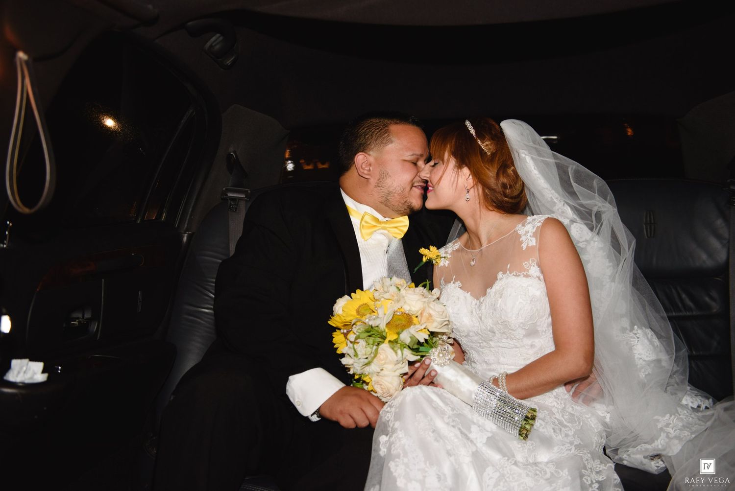 Bride and groom sharing a romantic moment in a limousine, bride holding yellow and white wedding bouquet.