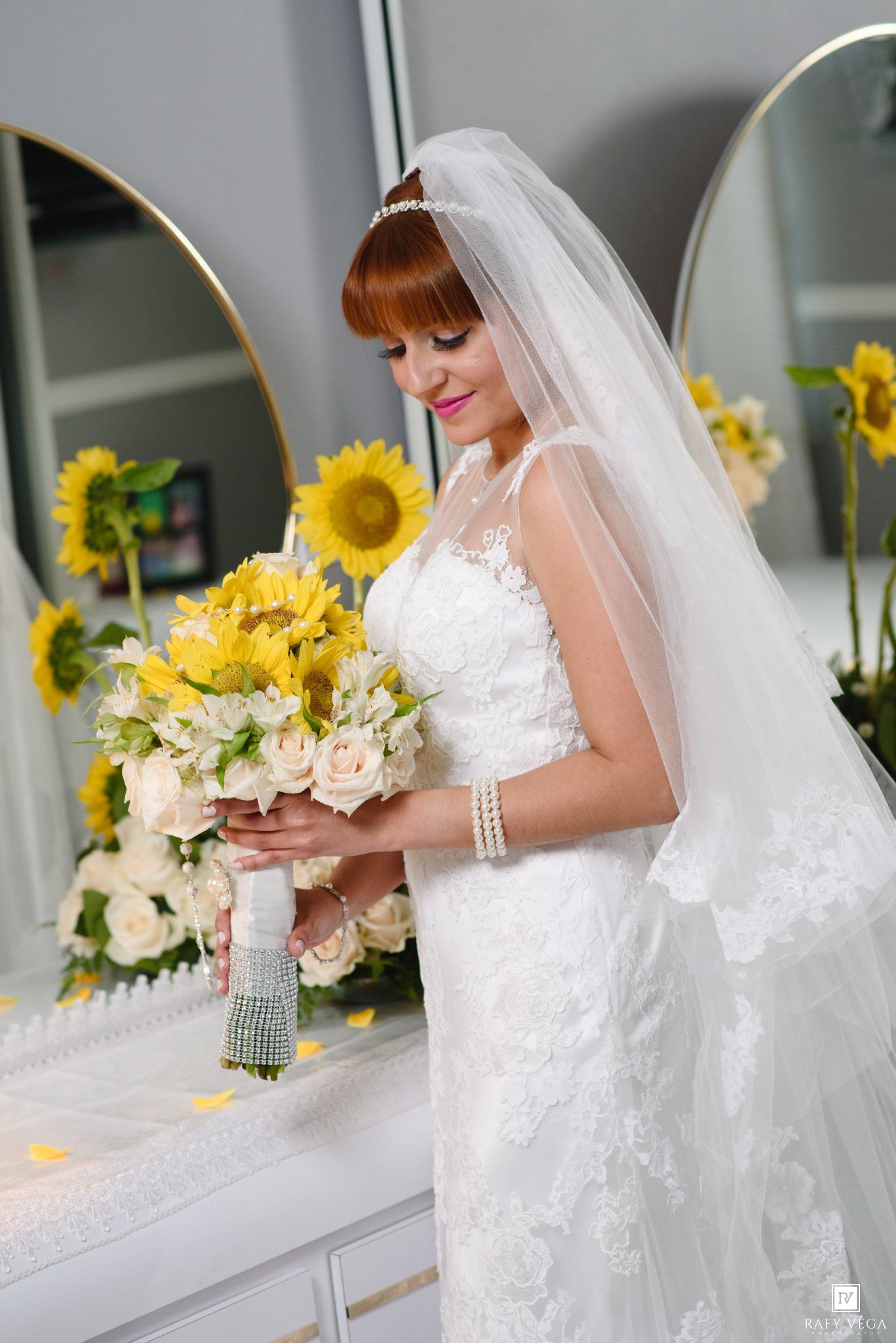 Bride in white lace gown and veil holds sunflower bouquet, smiling softly in a sunflower-decorated dressing room.