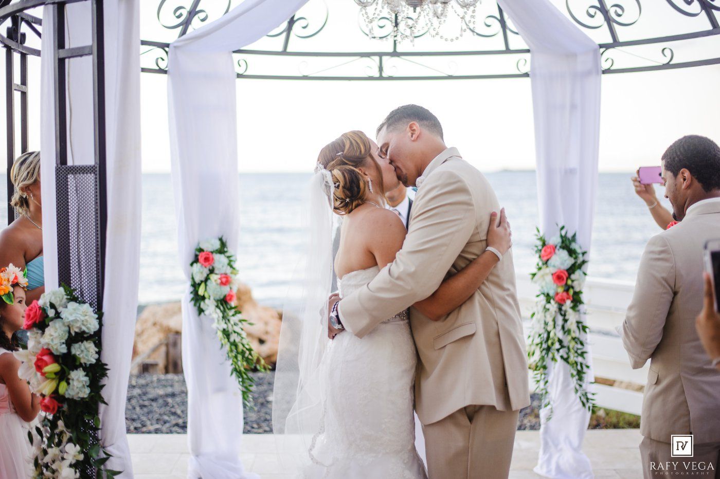 Newlyweds embrace in romantic first kiss under elegant iron gazebo decorated with white draping and tropical flowers by the ocean.