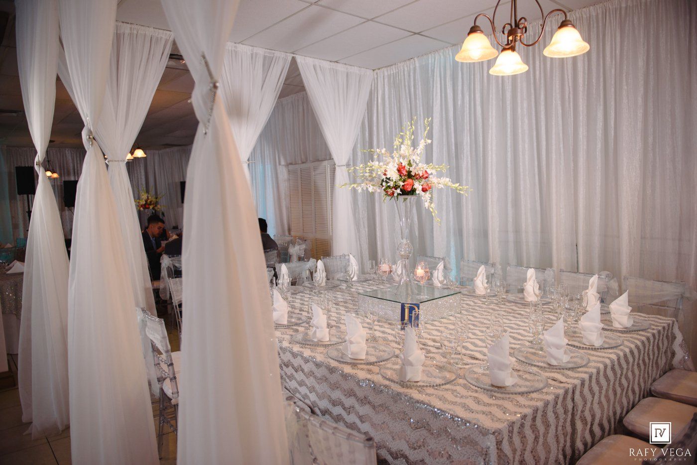 Elegant wedding reception venue with white draping, chevron tablecloth, floral centerpiece, and chandelier lighting.