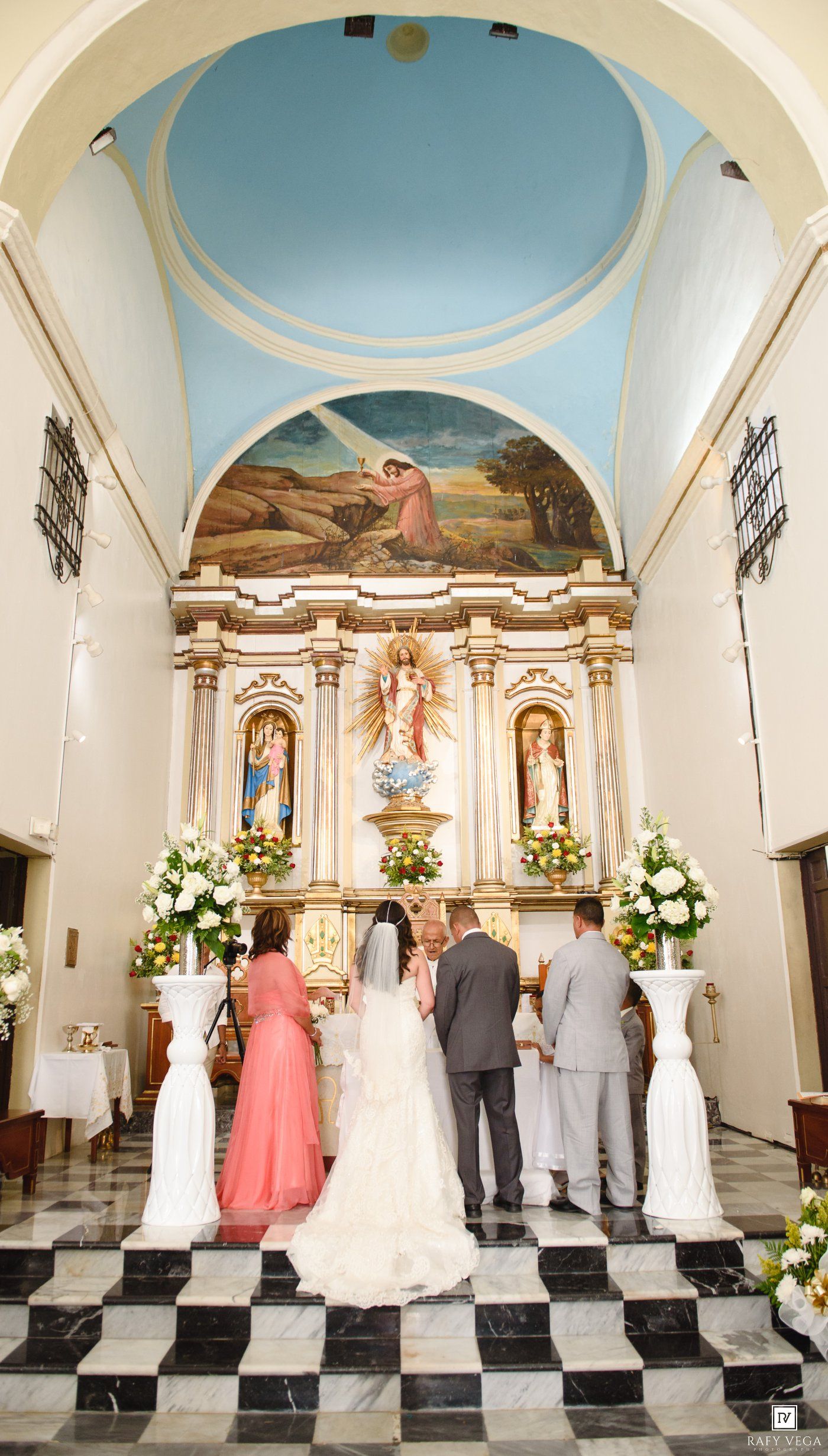 Wedding ceremony at ornate church altar with bride, groom, and attendants beneath blue dome and religious artwork.