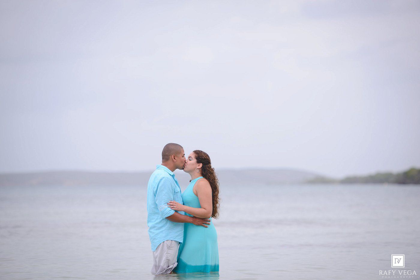 Couple sharing a romantic kiss on a tropical beach with calm ocean waters and overcast skies in the background.