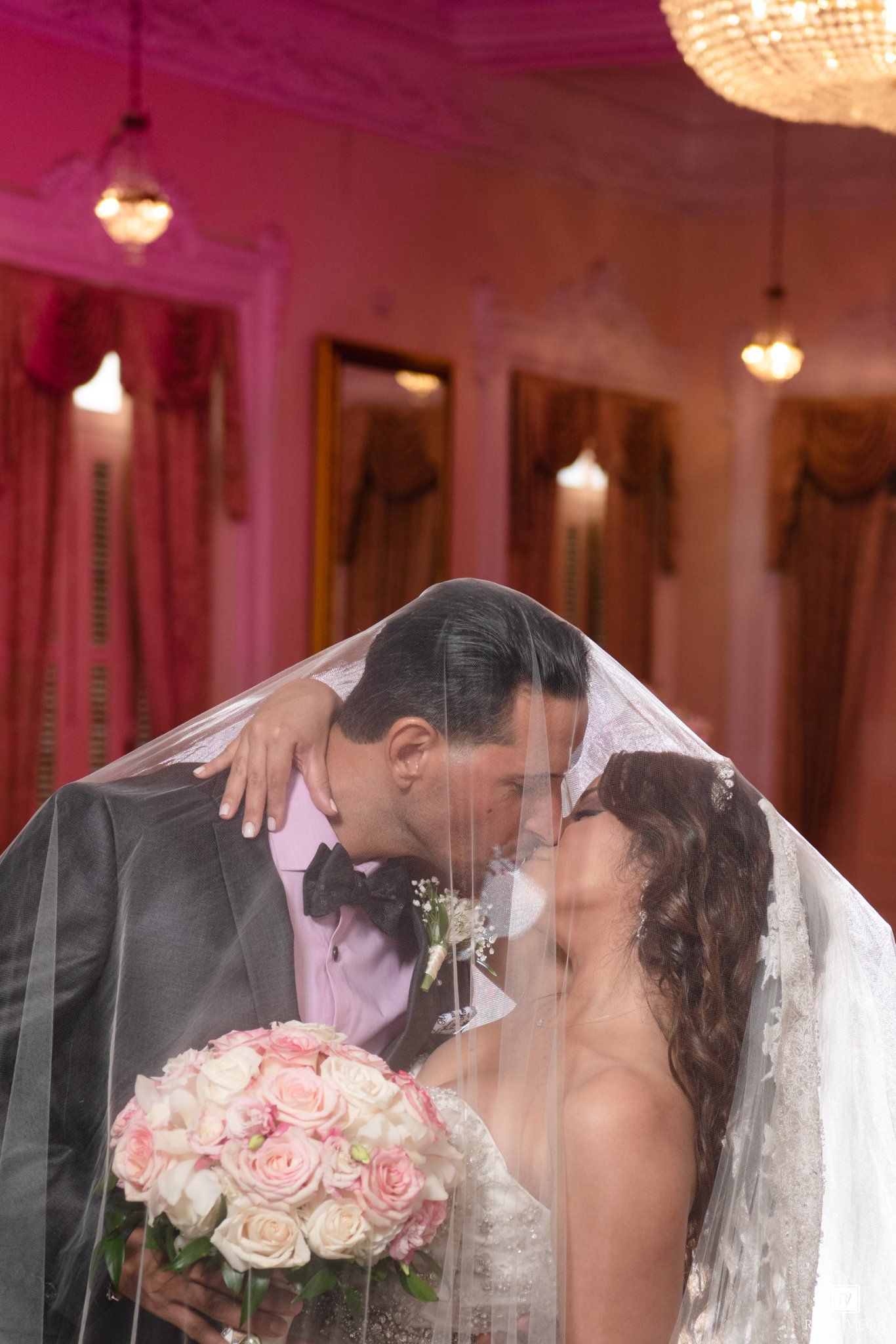 Bride and groom kissing under a white veil, bride holding pink roses bouquet in elegant pink ballroom.