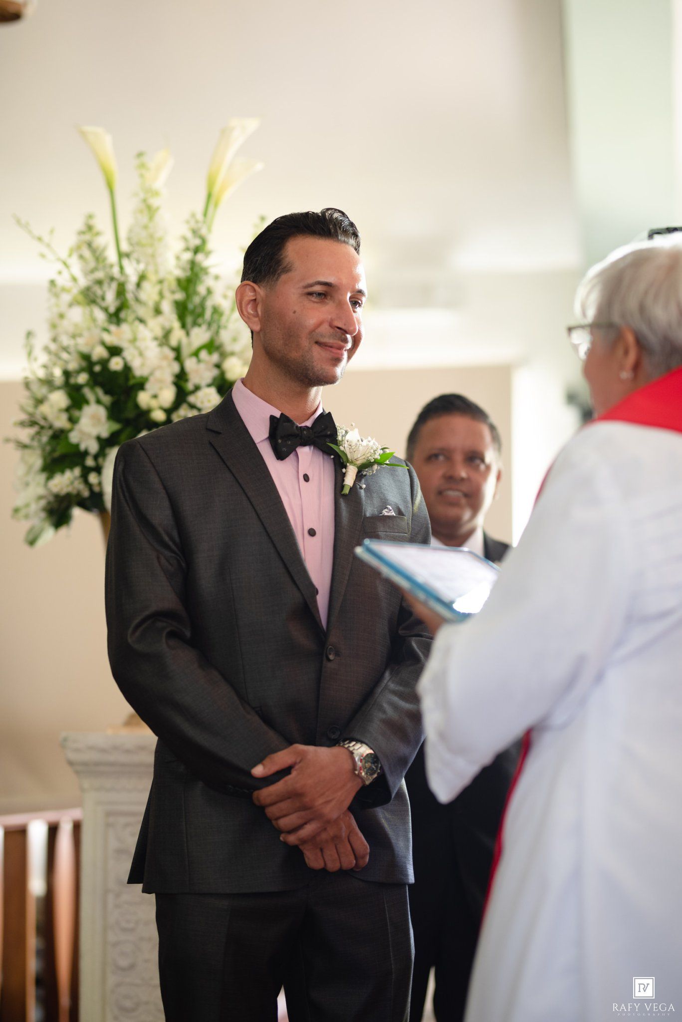Groom in gray tuxedo smiling at officiant during wedding ceremony in church with white floral arrangement.