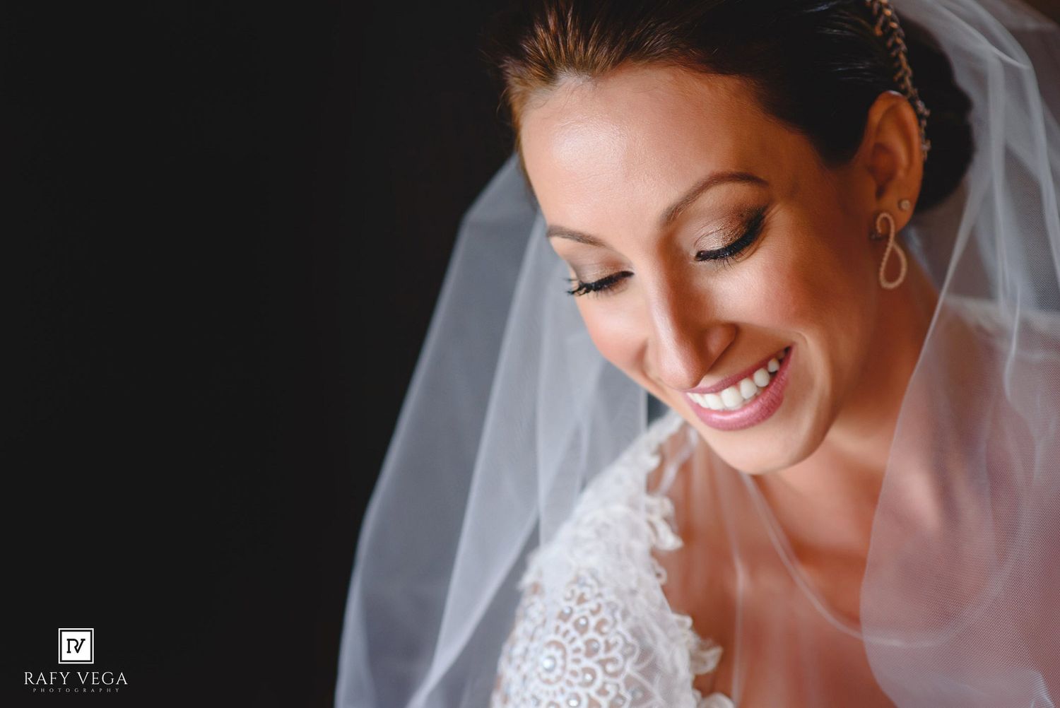 Smiling bride in white lace dress and veil with elegant makeup on dark background.