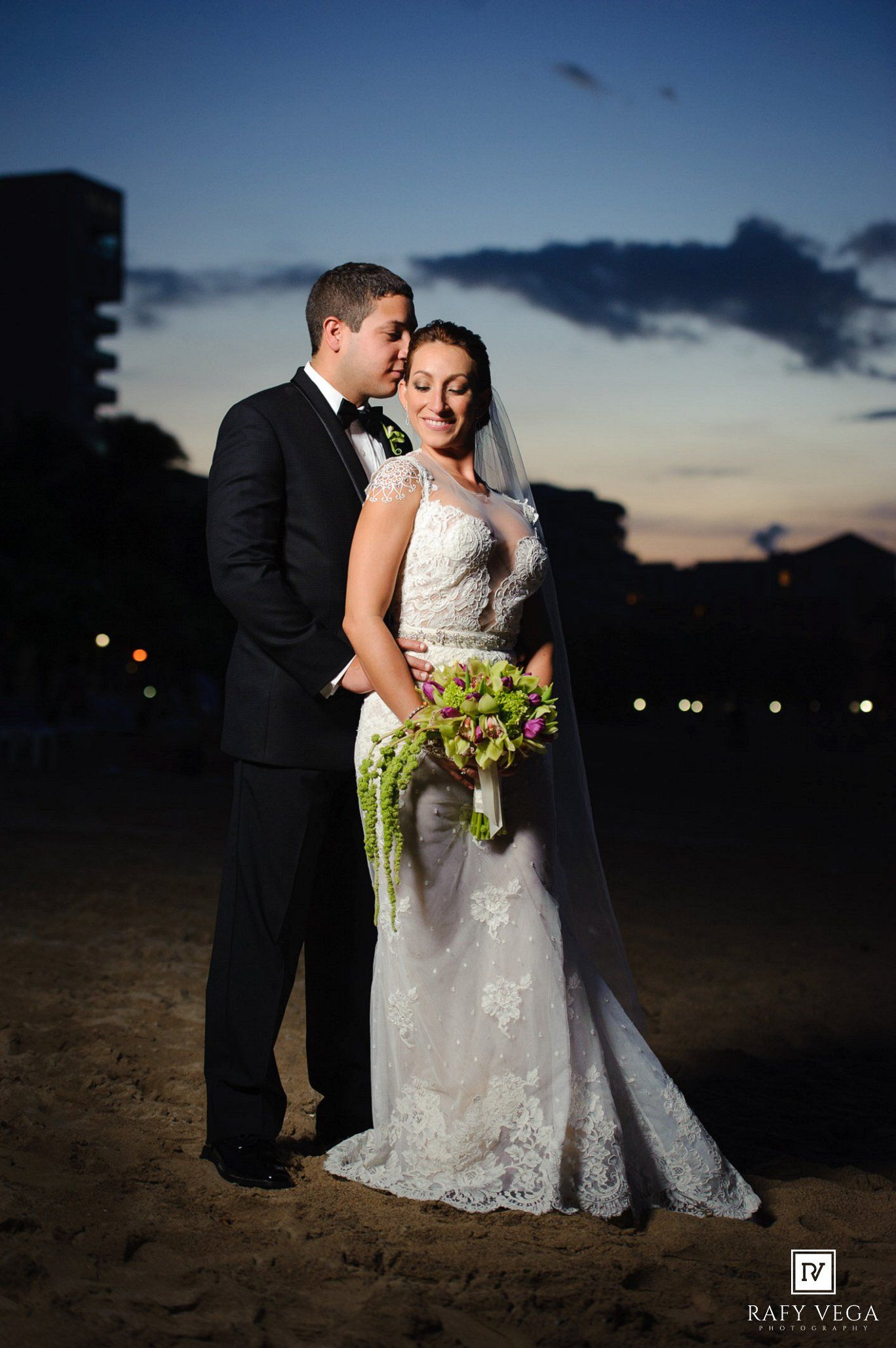 Bride and groom share a romantic beach moment at sunset, bride holding green floral bouquet in lace wedding gown.