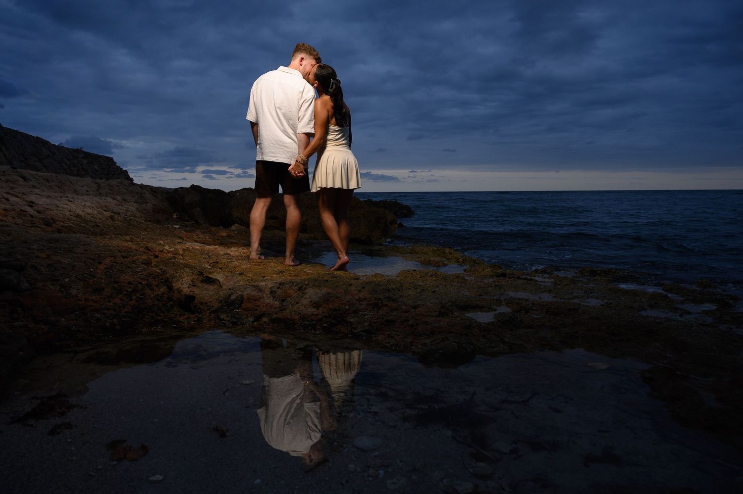 Romantic couple kissing at rocky shoreline at dusk, with their reflection visible in a tide pool below.