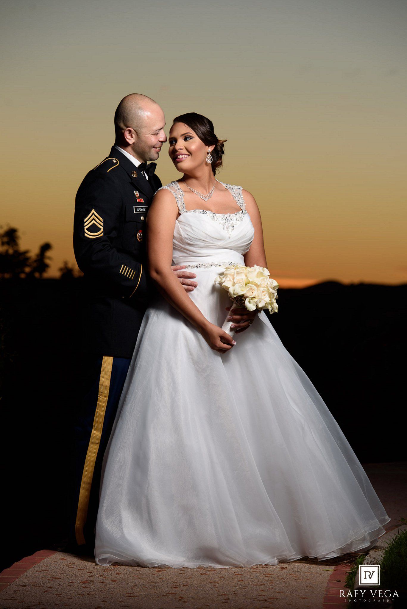 Military groom in dress uniform with bride in white ballgown holding bouquet at sunset wedding portrait.