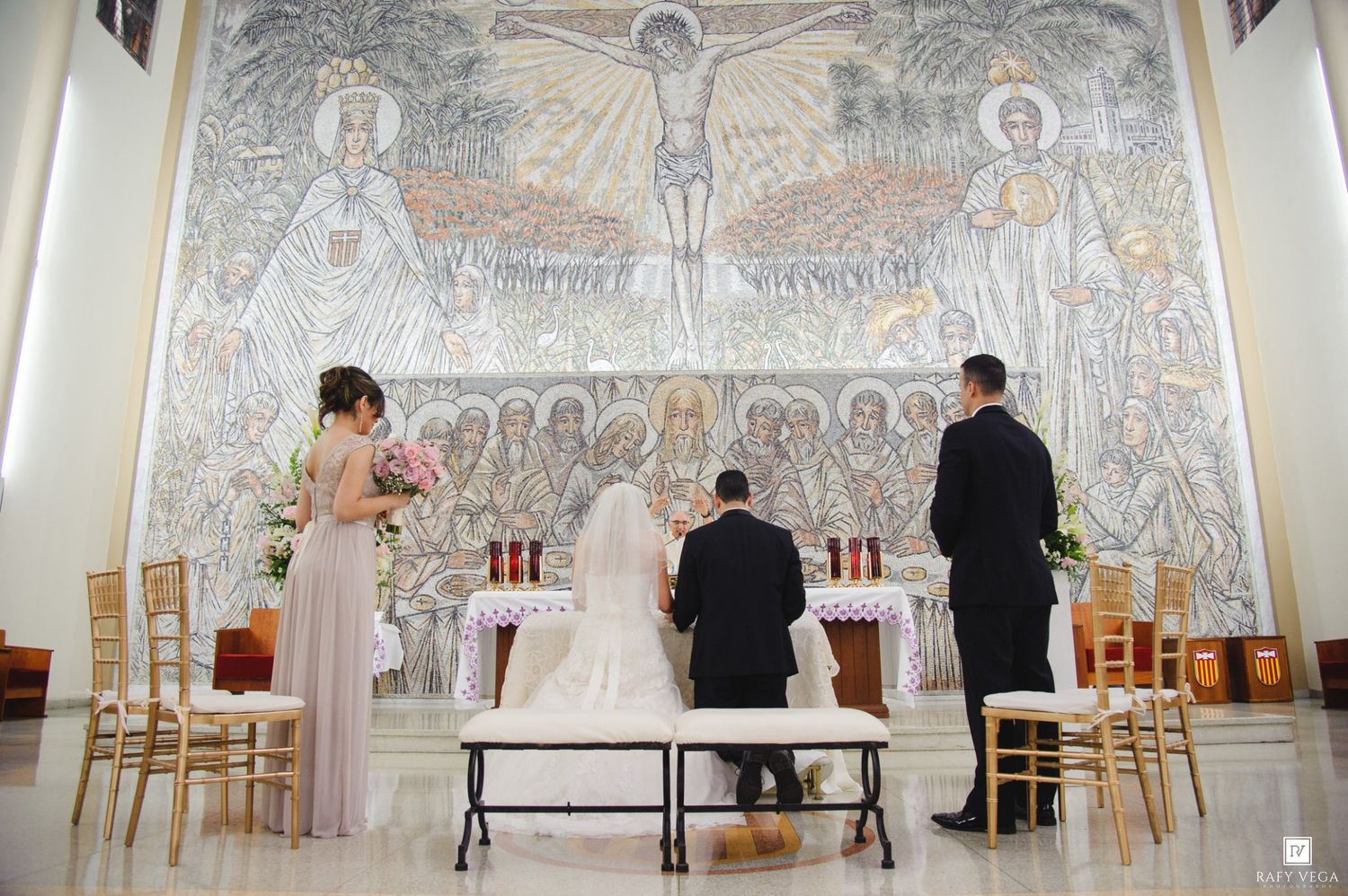 Wedding ceremony at Catholic church with couple kneeling at altar before large mosaic of crucifixion.