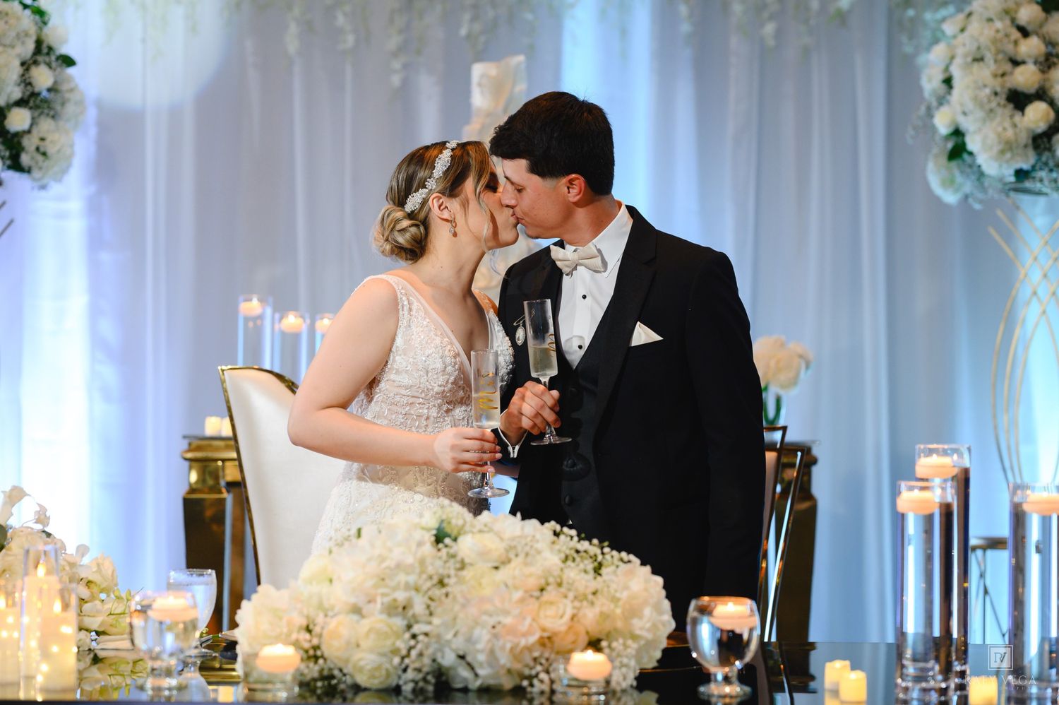 Bride and groom share a kiss while holding champagne glasses at elegant white floral wedding reception.