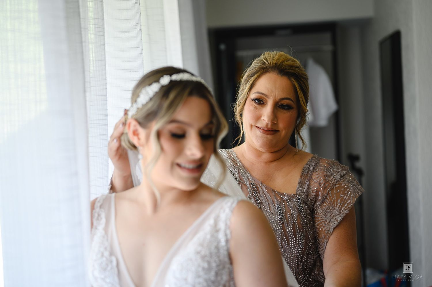 Bride adjusting her hair accessory while a smiling woman in a silver dress assists her during wedding preparations.
