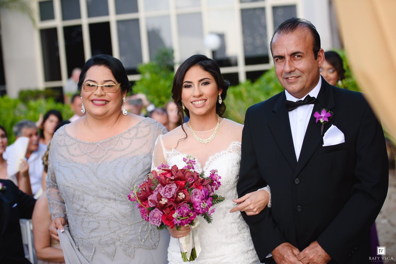 Bride in white gown holding pink bouquet poses with two guests at an elegant outdoor wedding ceremony.