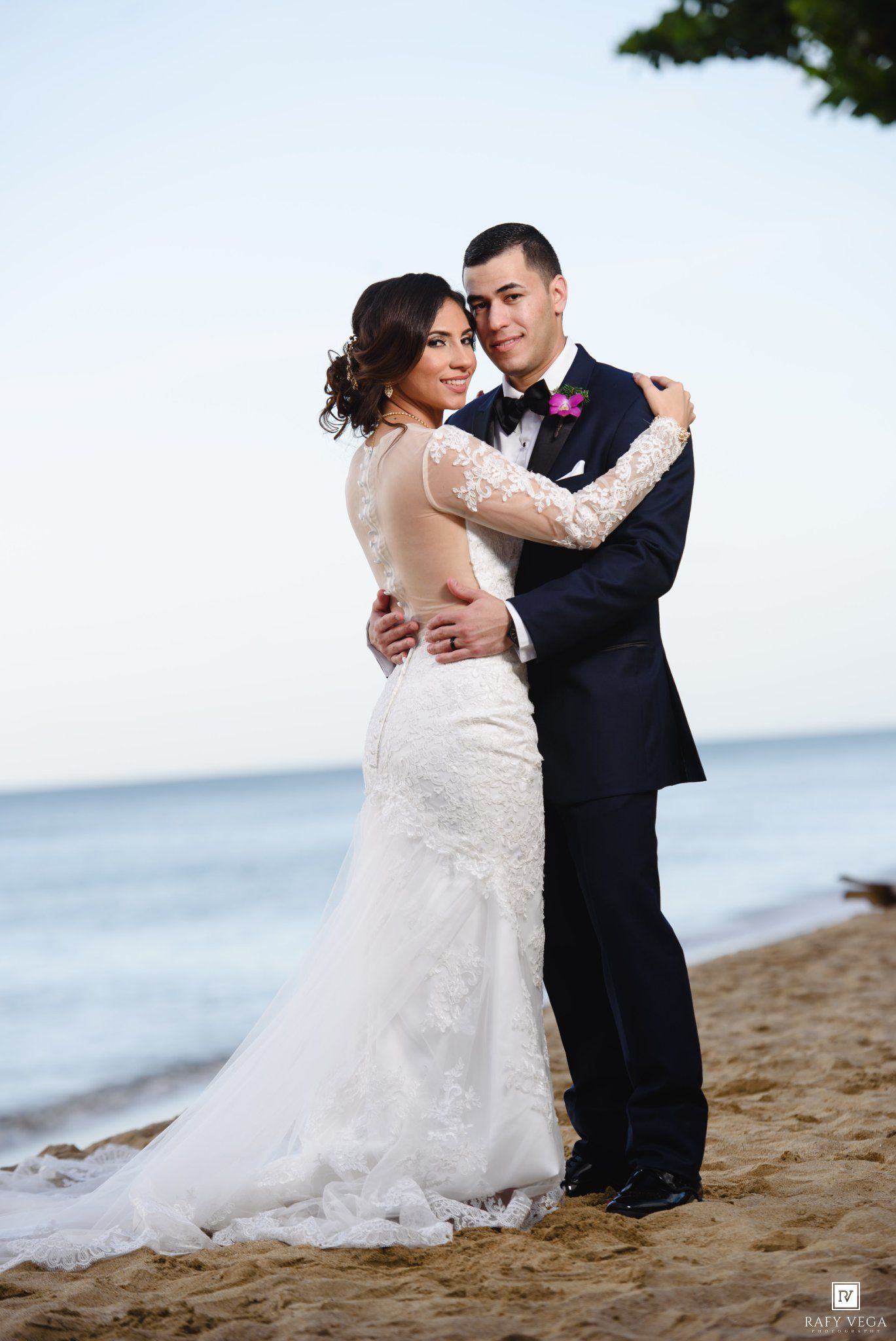 Bride and groom embrace on sandy beach, she in lace backless gown, he in navy tuxedo with pink boutonniere.