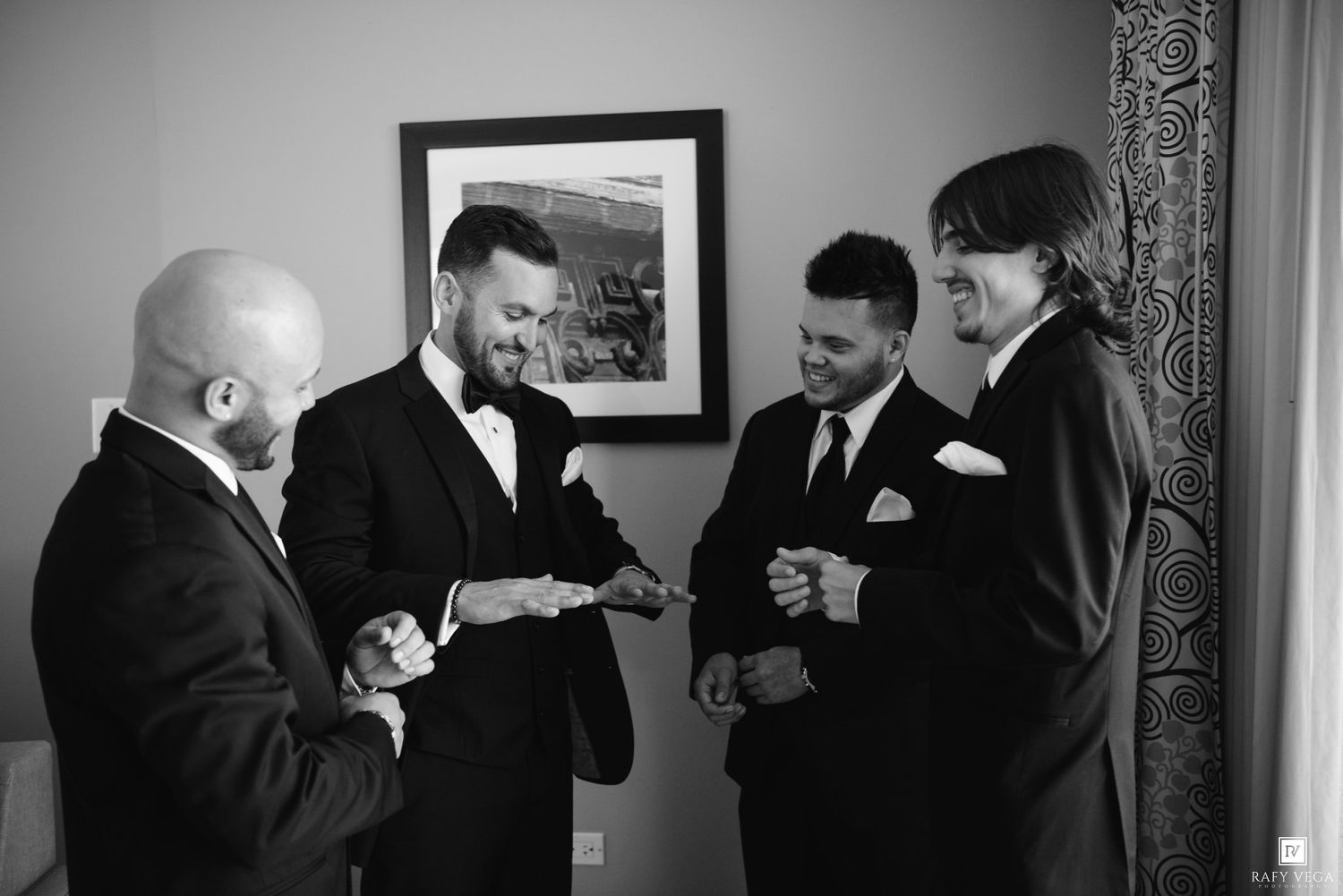 Groom and groomsmen laughing together in tuxedos before wedding ceremony in black and white photo.