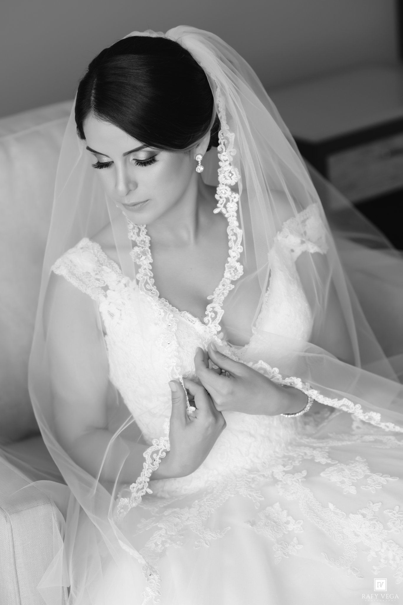 Black and white photo of a bride in lace wedding dress and veil, looking down while adjusting her gown.