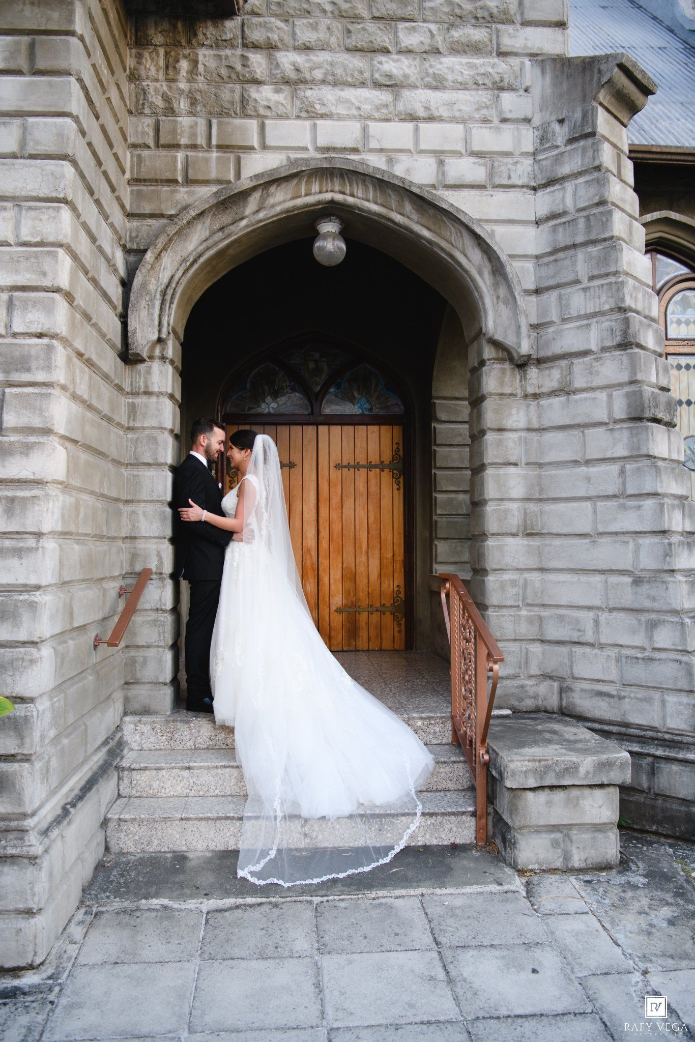 Bride and groom share a romantic kiss in a stone church archway, her white veil cascading down the steps.