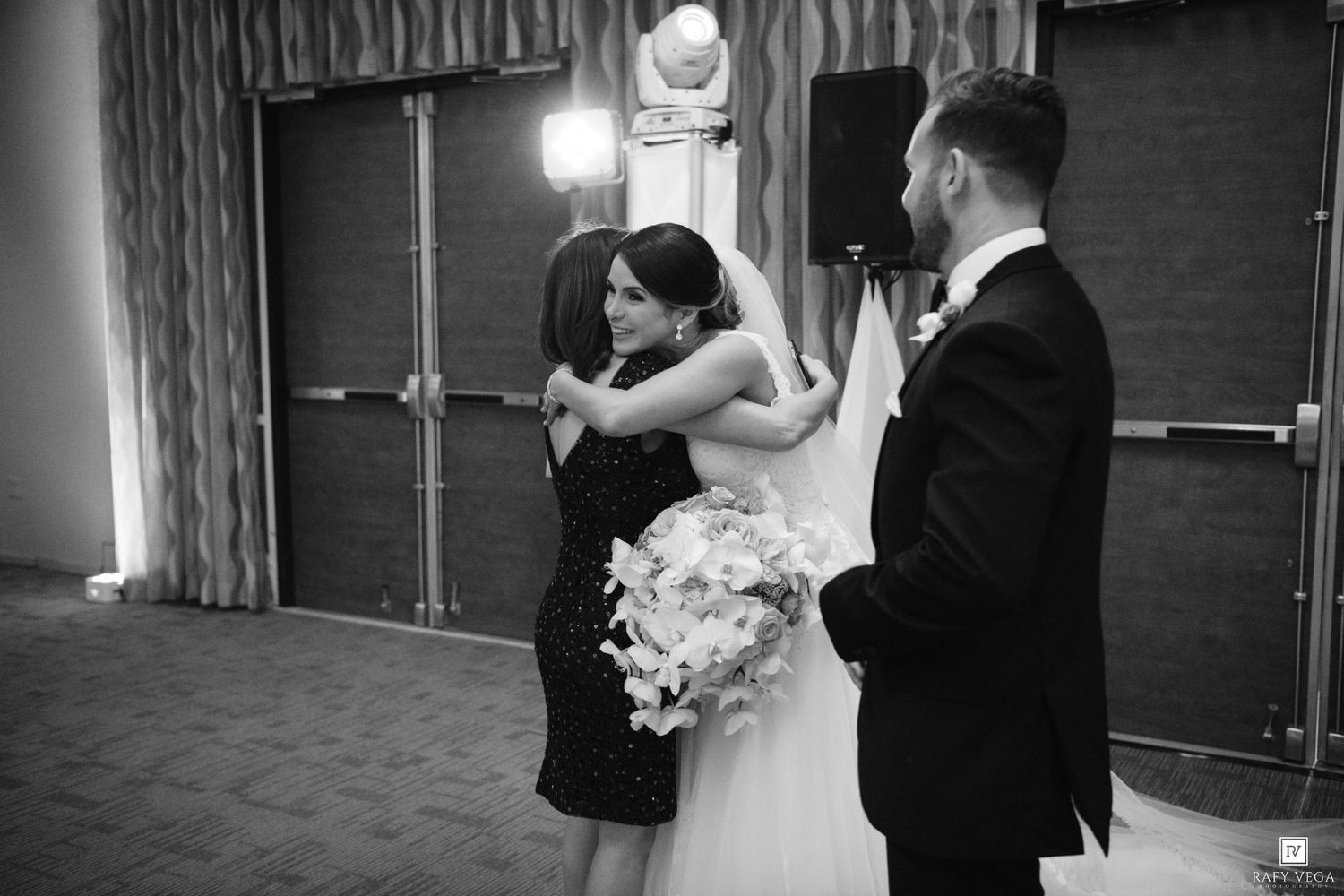 Bride and groom embrace a woman in black dress while holding a bouquet in a black and white wedding photo.