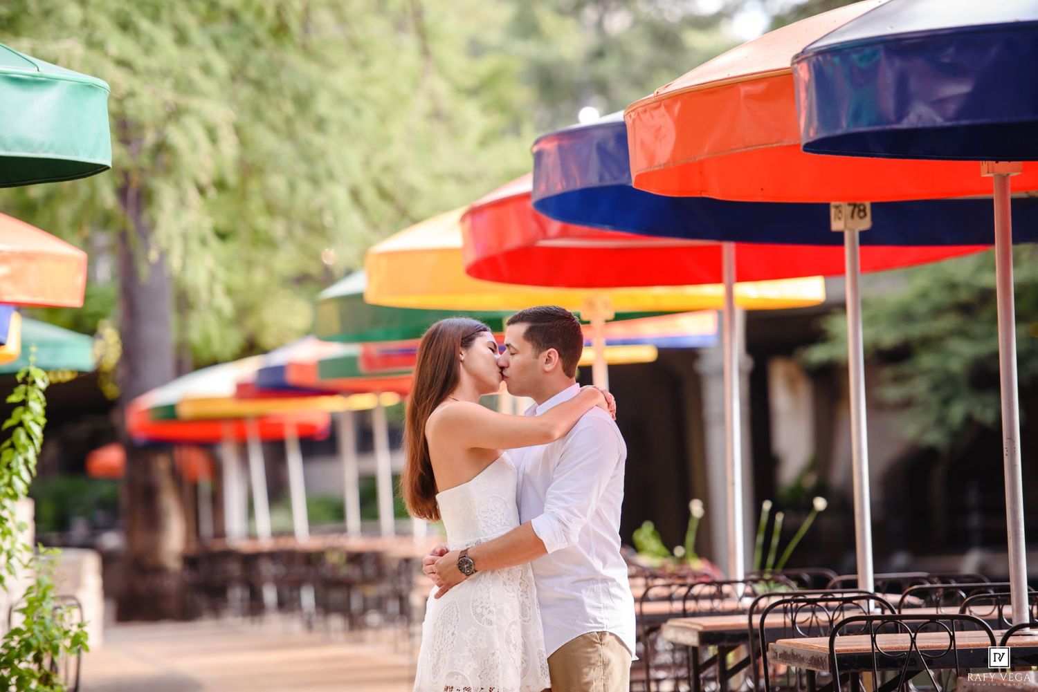 Couple sharing a romantic kiss under colorful outdoor umbrellas at a vibrant cafe patio.