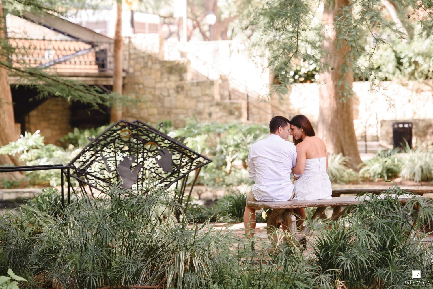 Couple sharing a tender kiss while seated on a bench surrounded by lush garden greenery.