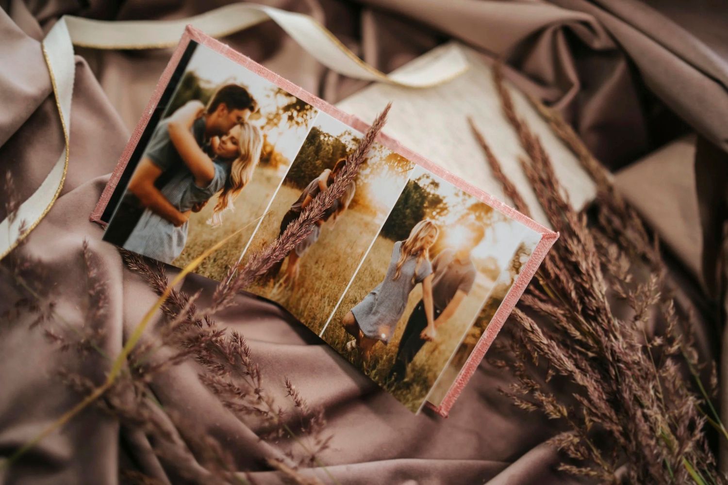 A romantic photo book with couple portraits rests on draped mauve fabric surrounded by dried pampas grass.