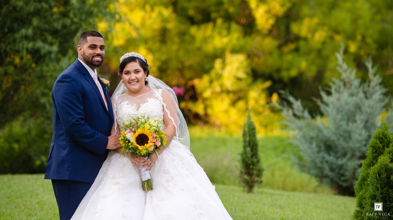 Bride and groom smiling outdoors in lush garden, bride holding sunflower bouquet in white gown, groom in navy suit.