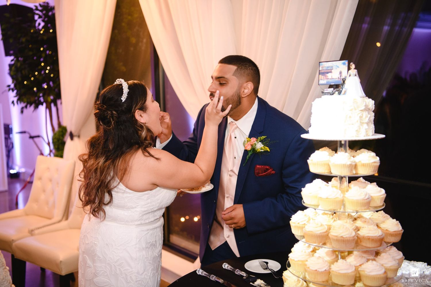 Bride feeds wedding cake to groom in blue suit at elegant reception with cupcake tower display.