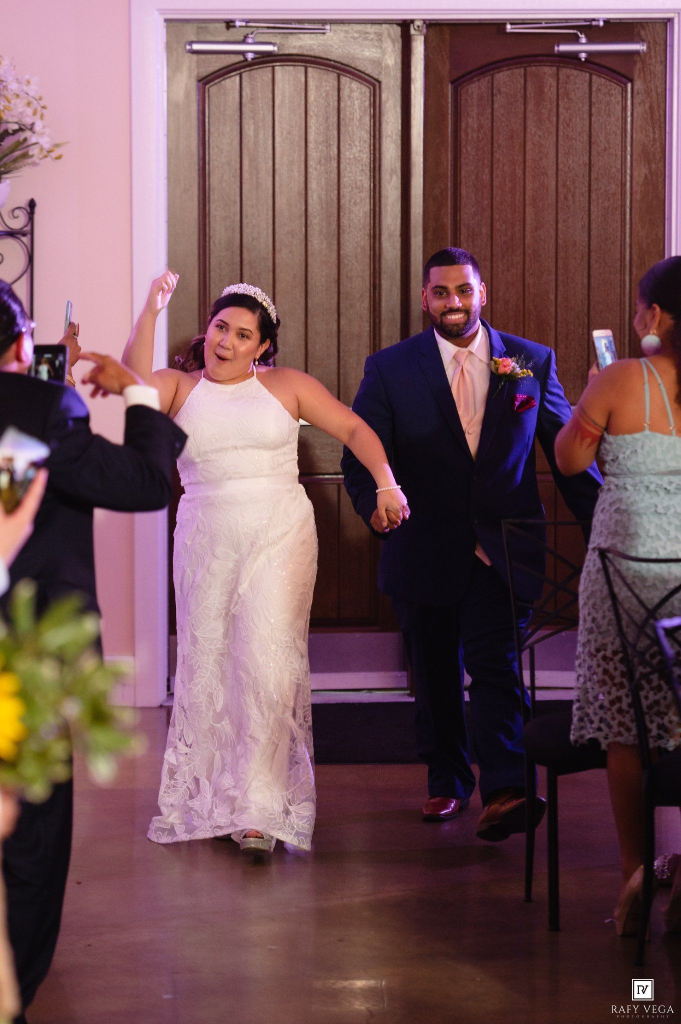 Bride in white halter gown and groom in navy suit make grand entrance at wedding reception, holding hands and cheering.