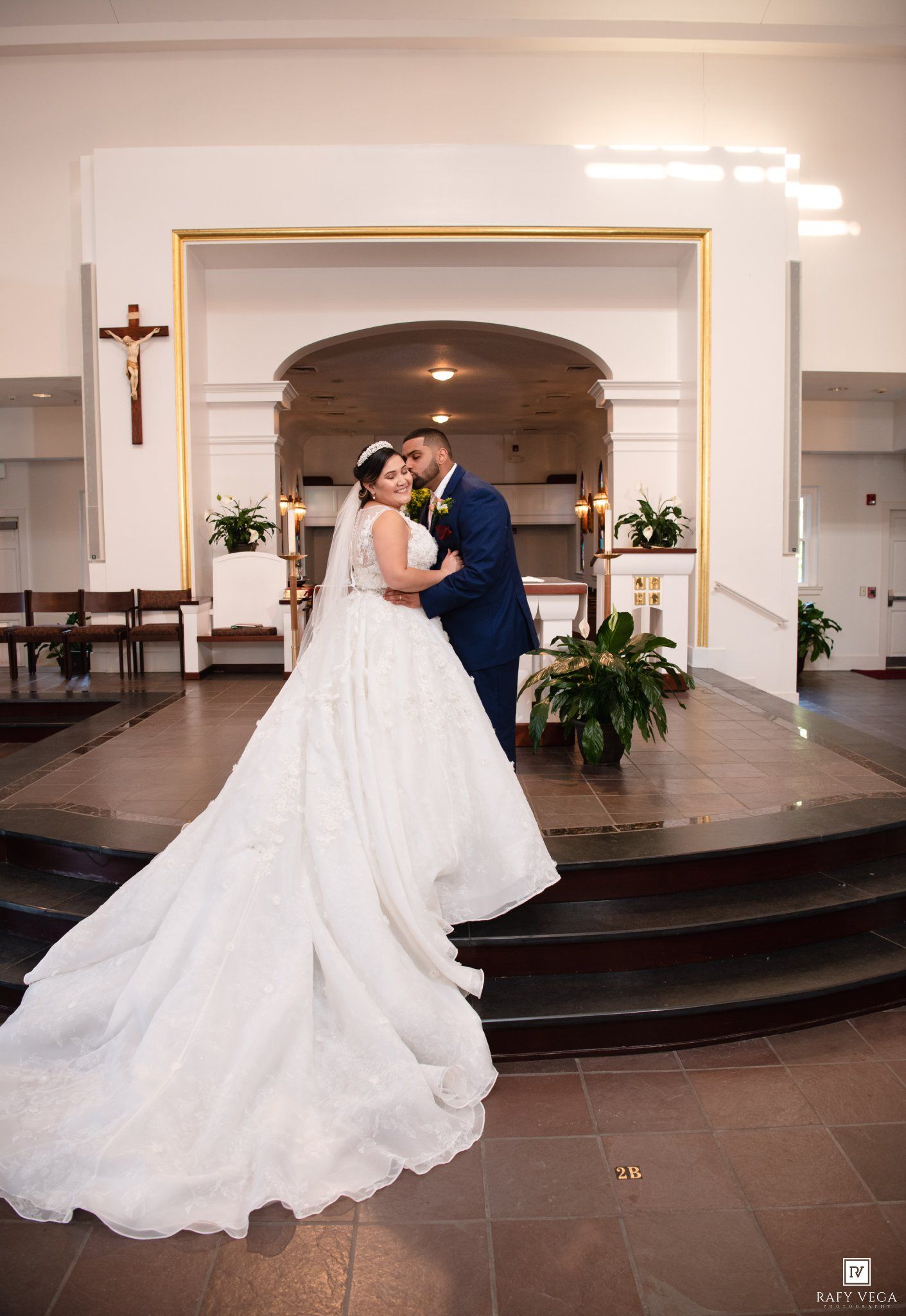 Bride and groom share a romantic embrace at church altar, bride wearing elegant white ball gown with long train.