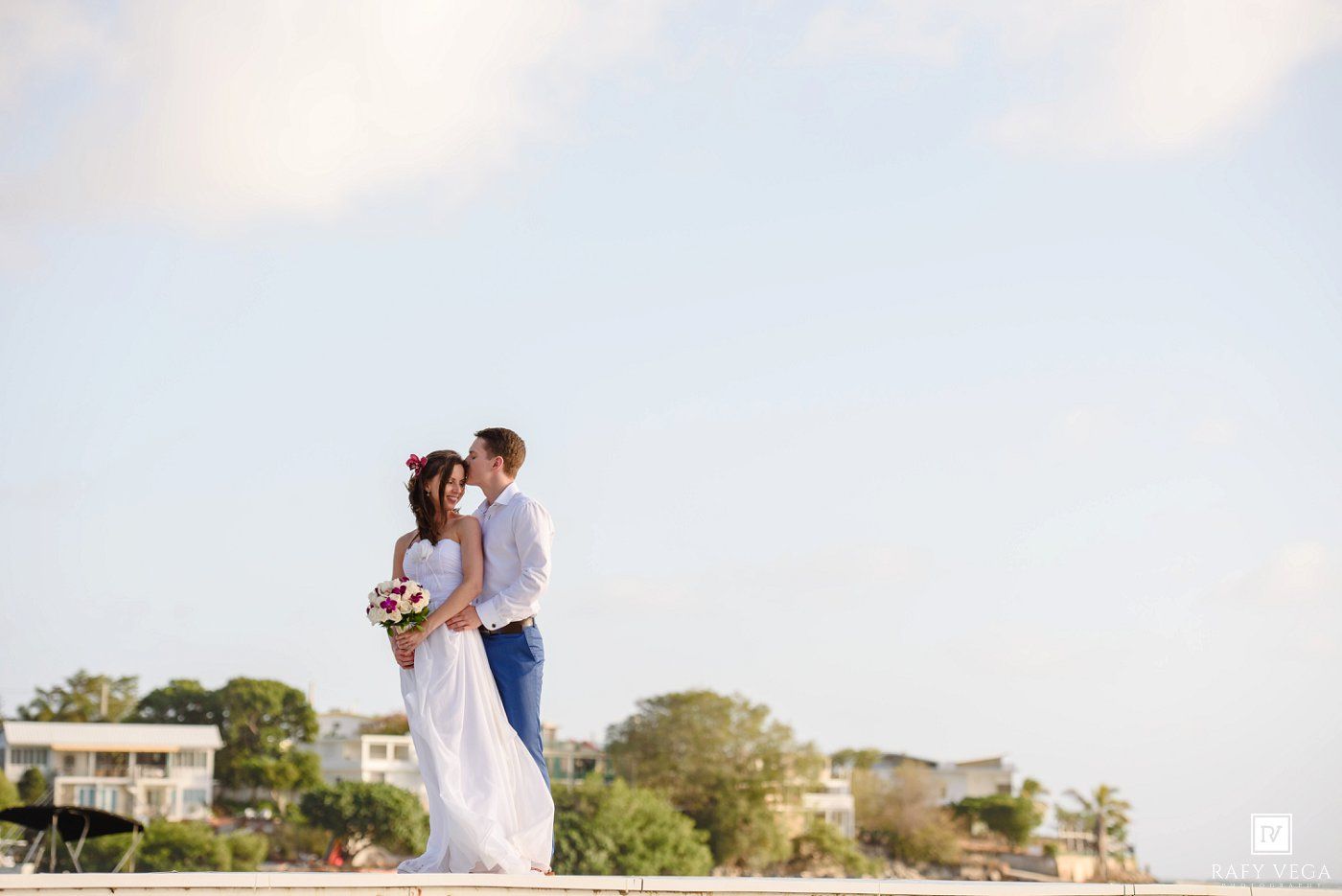 A bride and groom share a romantic embrace outdoors with a scenic coastal backdrop on their wedding day.