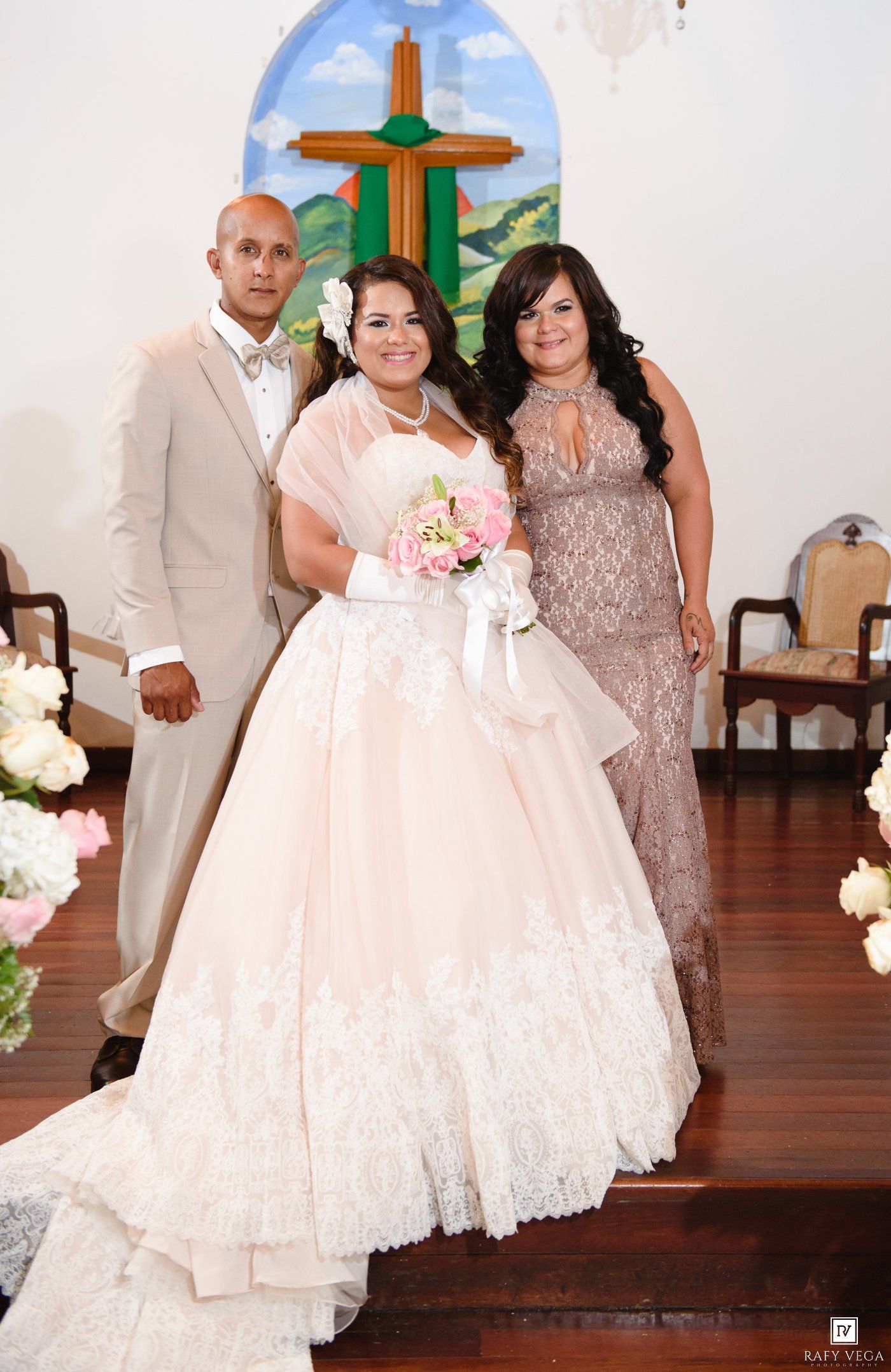 Bride in blush ball gown with lace holds pink bouquet, flanked by two guests in a church with wooden cross.