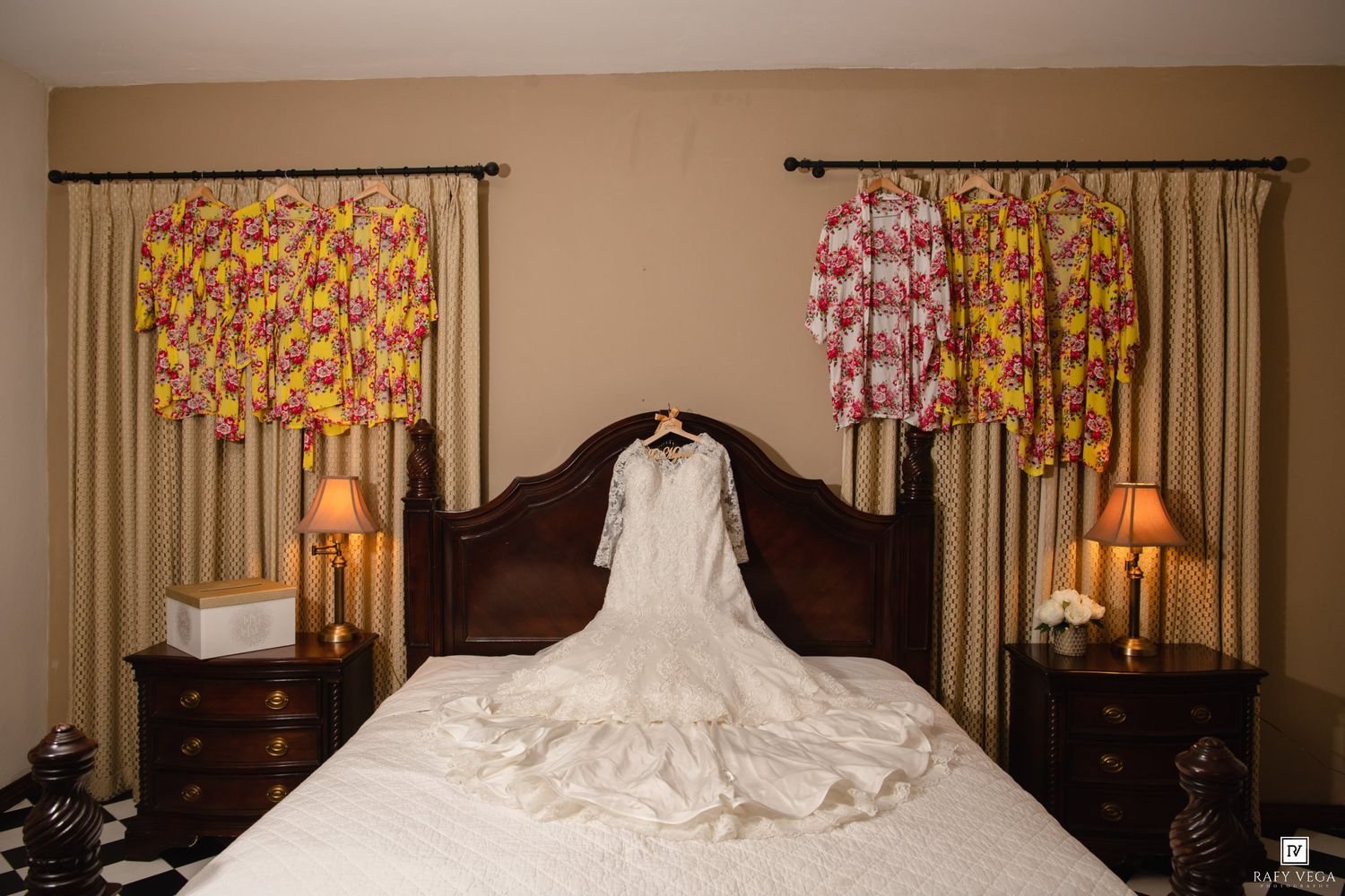 White wedding gown hanging over bed with colorful floral robes on curtain rods in elegant hotel bedroom.