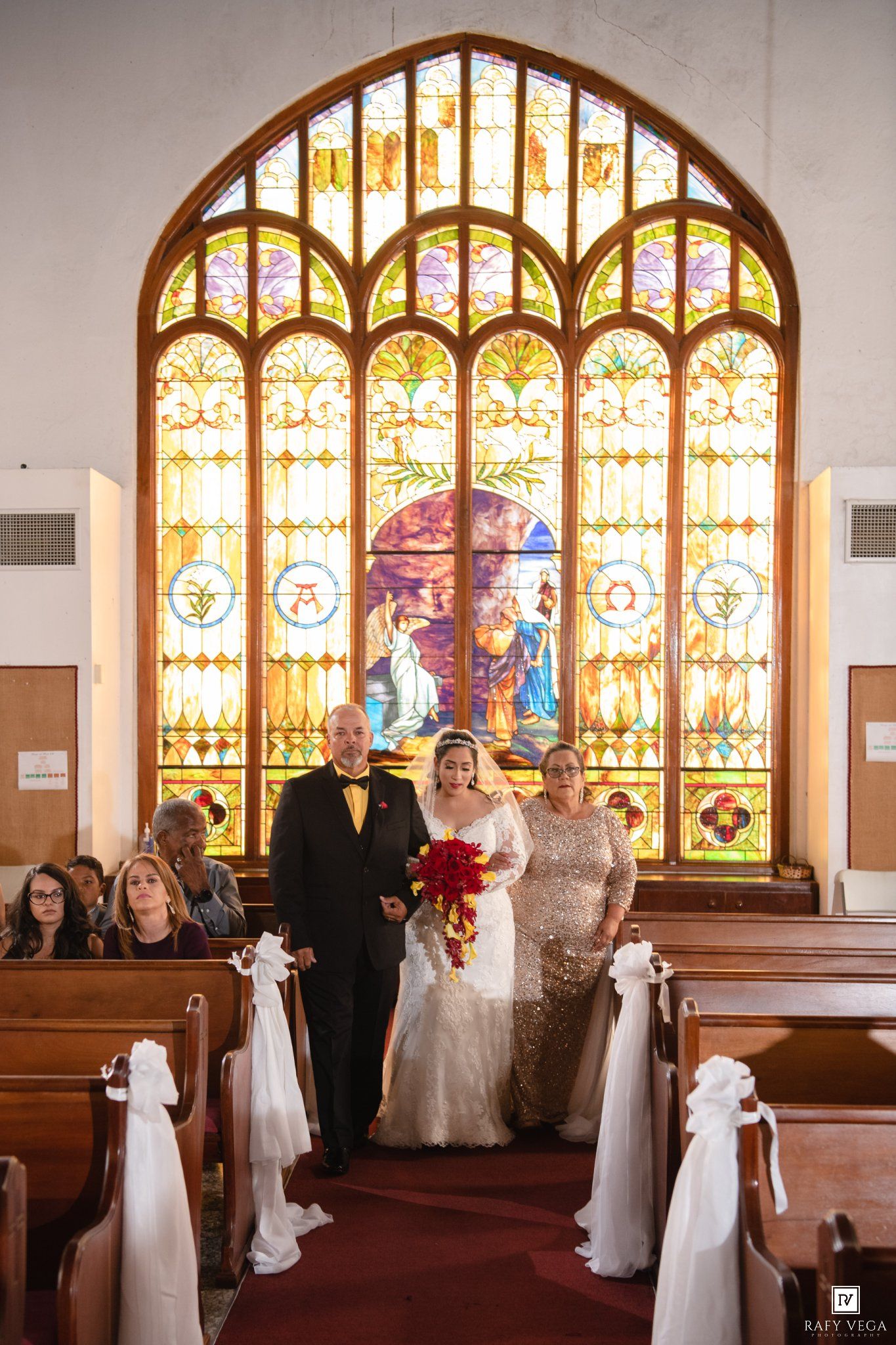 Bride walks down church aisle with parents before stunning stained glass window, holding red bouquet.