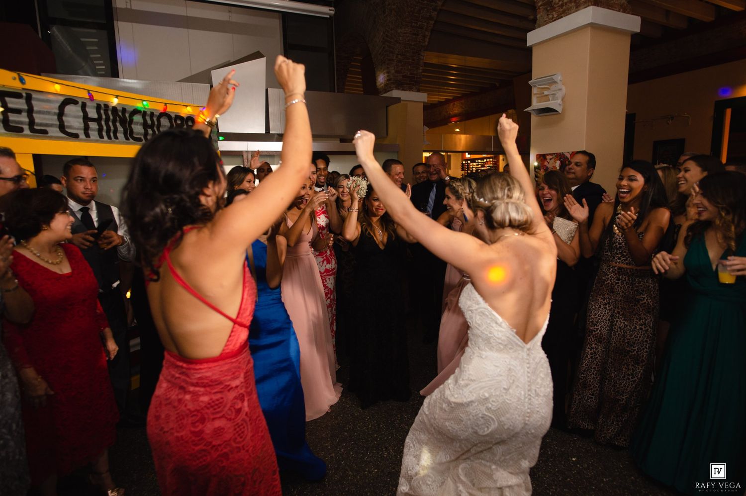 Bride in white gown and guest in red dress dancing with arms raised at a lively wedding reception party.