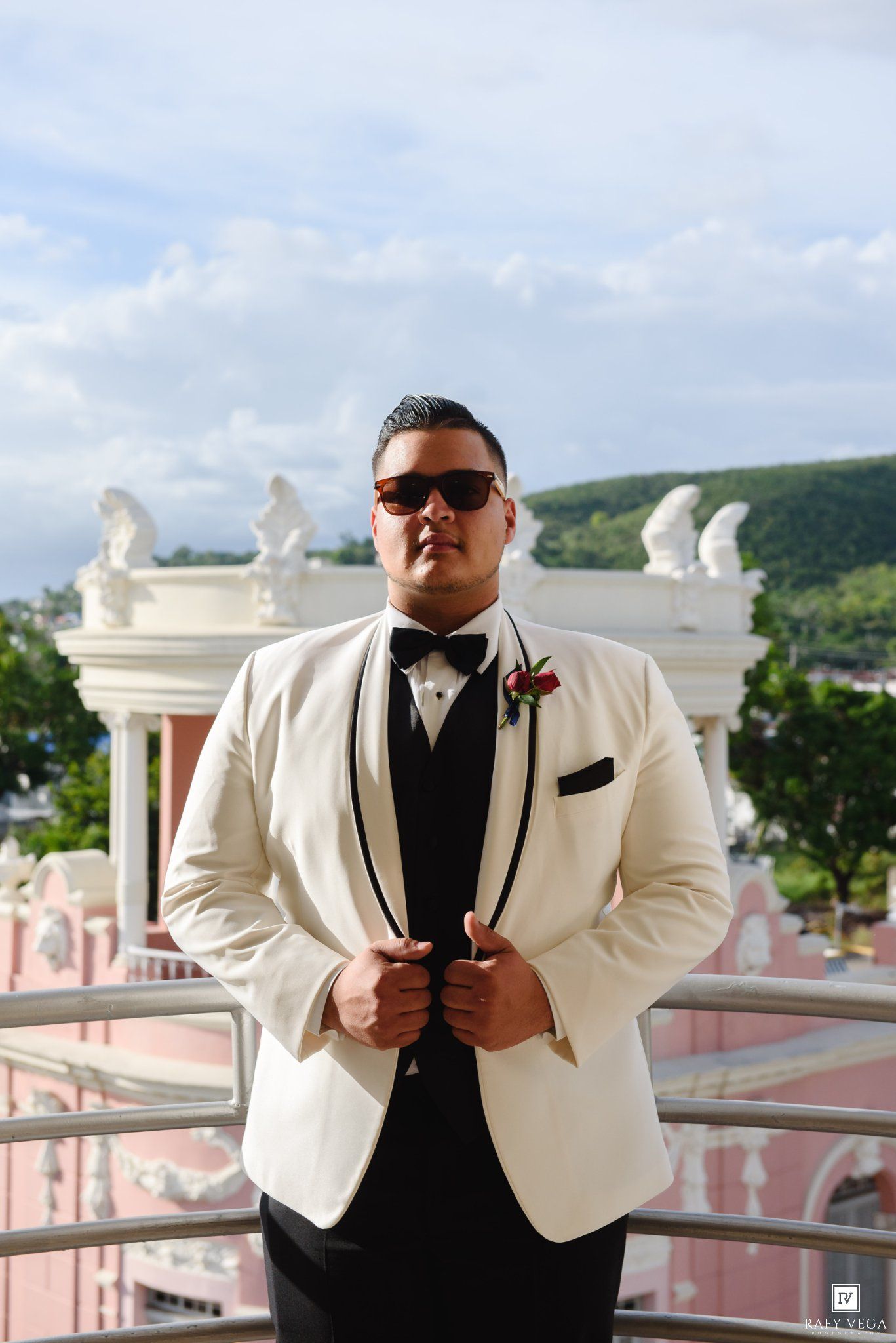 Groom in white tuxedo jacket, black bow tie, and sunglasses poses on balcony with pink building and green hills behind.